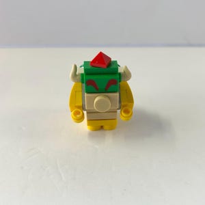 Peut inclure: Une petite figurine Bowser color&eacute;e construite &agrave; partir de briques de jouets embo&icirc;tables. La figurine pr&eacute;sente un corps jaune, une carapace verte et un joyau rouge sur la t&ecirc;te. Elle a des cornes blanches et un visage beige avec des sourcils rouges.