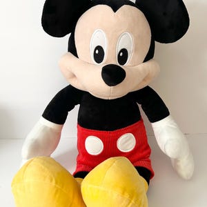 May include: A plush Mickey Mouse toy featuring large black ears, a tan face with white eyes, and a black nose. The character wears red corduroy shorts with two white circular buttons and large yellow shoes, with white gloves on its hands.