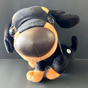 May include: A plush dog toy with a large, brown nose and big, round eyes. The toy has black fur with tan accents on the ears, paws, and around the face. The dog is in a sitting position.