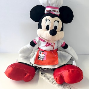 Op de afbeelding: Een pluche Minnie Mouse pop verkleed als chef-kok, met een witte koksmuts met roze rand en de tekst "Chef Mickey's". Ze draagt een wit koksuniform met een rode schort met een cupcake-ontwerp. Haar grote rode schoenen en zwarte oren zijn prominent aanwezig.