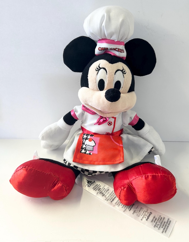 Op de afbeelding: Een pluche Minnie Mouse pop verkleed als chef-kok, met een witte koksmuts met roze rand en de tekst "Chef Mickey's". Ze draagt een wit koksuniform met een rode schort met een cupcake-ontwerp. Haar grote rode schoenen en zwarte oren zijn prominent aanwezig.
