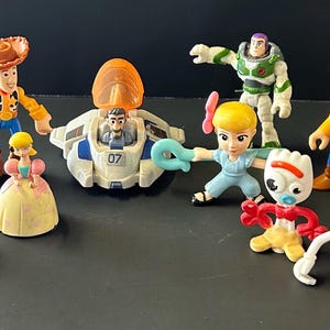Op de afbeelding: Een verzameling plastic Toy Story-figuren, waaronder Woody, Buzz Lightyear, Jessie, Bo Peep, Forky, Mr. Potato Head, Bullseye, twee groene aliens, een kleine prinsesfiguur en een wit met blauw ruimteschip met een oranje koepel.