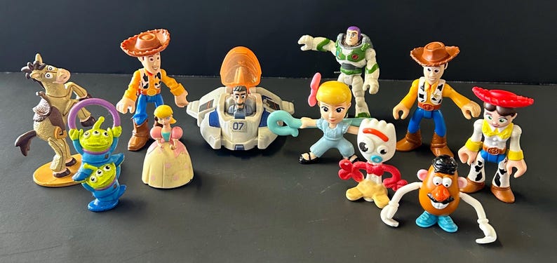 Op de afbeelding: Een verzameling plastic Toy Story-figuren, waaronder Woody, Buzz Lightyear, Jessie, Bo Peep, Forky, Mr. Potato Head, Bullseye, twee groene aliens, een kleine prinsesfiguur en een wit met blauw ruimteschip met een oranje koepel.