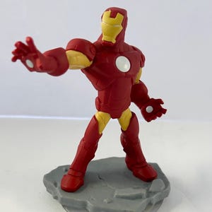 May include: A red and yellow Iron Man action figure, posed with an outstretched arm. The figure has a red suit with yellow accents on the joints and helmet. It stands on a gray, textured base.