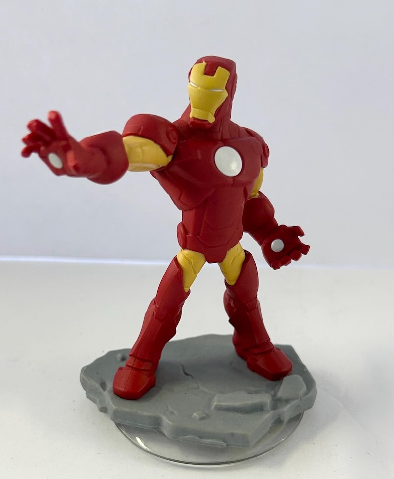 May include: A red and yellow Iron Man action figure, posed with an outstretched arm. The figure has a red suit with yellow accents on the joints and helmet. It stands on a gray, textured base.
