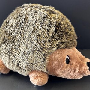 May include: A plush hedgehog toy featuring a rounded body covered in long, shaggy brown and tan faux fur. The face, underbelly, and small paws are made of smooth, light brown fabric. It has a small black nose and dark, round eyes.