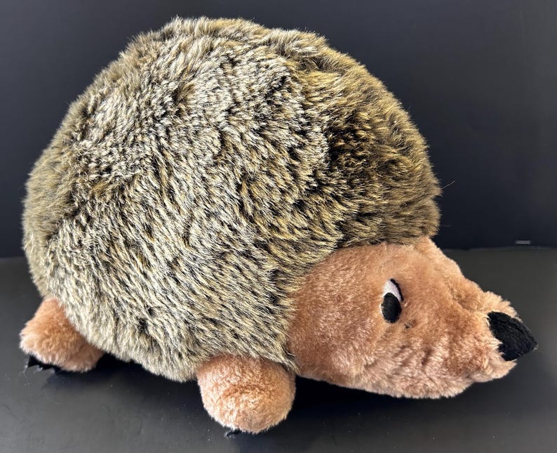 May include: A plush hedgehog toy featuring a rounded body covered in long, shaggy brown and tan faux fur. The face, underbelly, and small paws are made of smooth, light brown fabric. It has a small black nose and dark, round eyes.