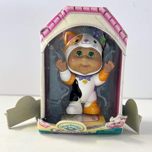 May include: A Cabbage Patch Kids figurine in a cat costume, displayed in a house-shaped box. The doll has a white, orange, and black outfit with a matching cat hood. The box is pink and white.