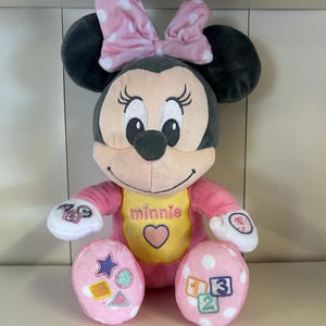May include: A plush Minnie Mouse doll with a pink bow and polka dots. The doll has a pink outfit, a yellow bib with the name "Minnie" and a heart, and pink feet with shapes and numbers. The doll is designed for children.