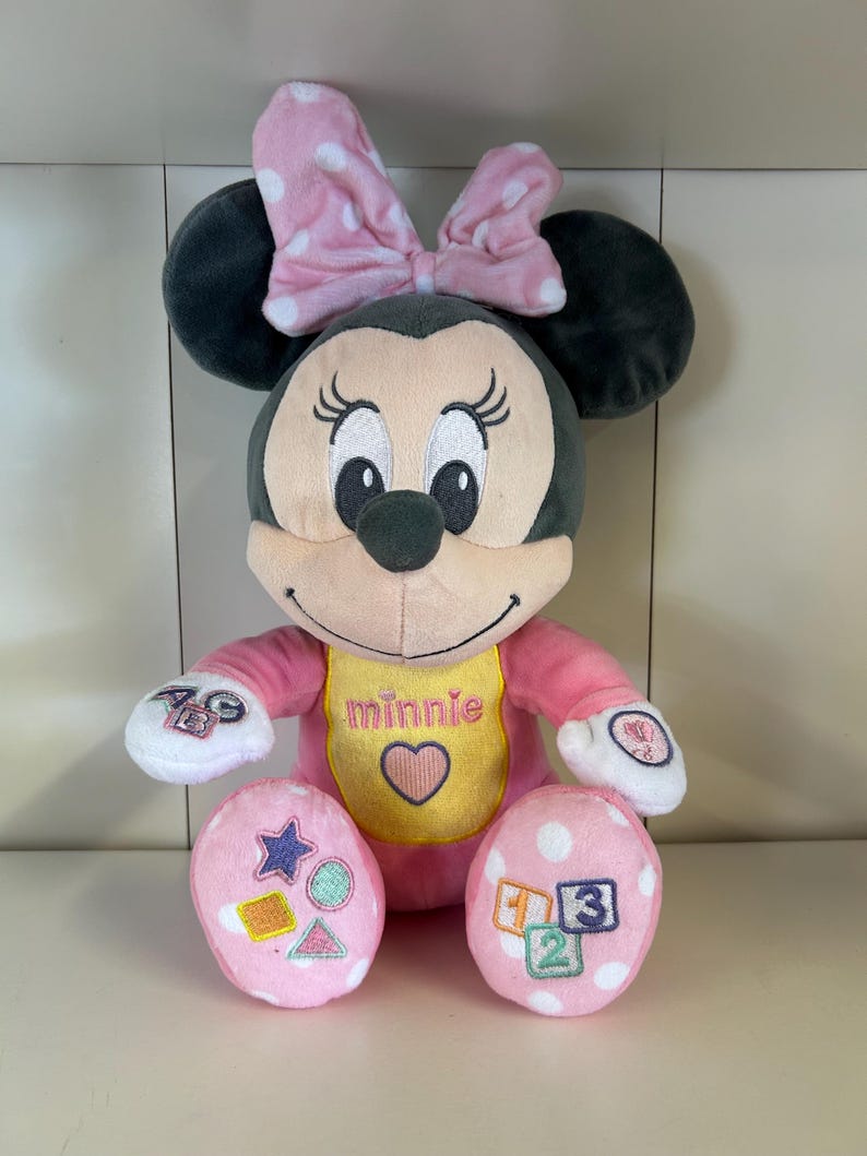 May include: A plush Minnie Mouse doll with a pink bow and polka dots. The doll has a pink outfit, a yellow bib with the name "Minnie" and a heart, and pink feet with shapes and numbers. The doll is designed for children.