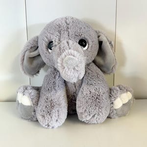 May include: A plush gray elephant toy with large ears, a trunk, and white accents on its feet. The stuffed animal has big black eyes and is sitting upright. The elephant is made of soft, fluffy material.