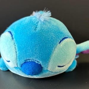 K&ouml;nnte beinhalten: Ein Pl&uuml;schtier in Form einer blauen Alien-Figur mit rundem K&ouml;rper, geschlossenen Augen, einer dunkelblauen Nase und gro&szlig;en Ohren mit violetten Innenseiten. Der weiche Stoff ist hellblau und eine kleine hellblaue Haarstr&auml;hne sitzt oben in der Mitte.