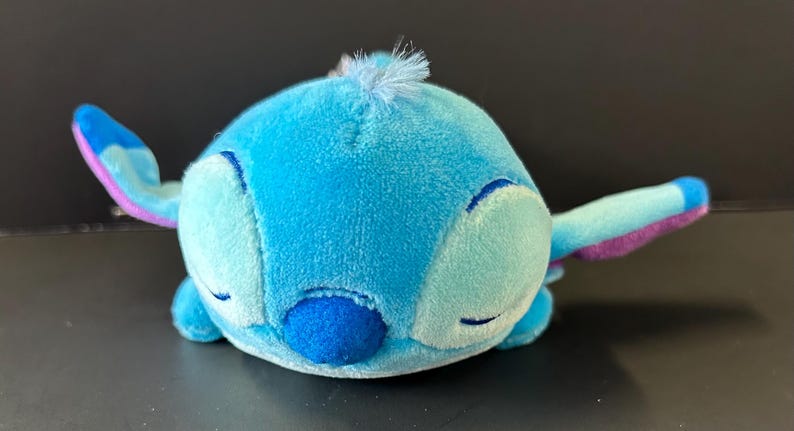 K&ouml;nnte beinhalten: Ein Pl&uuml;schtier in Form einer blauen Alien-Figur mit rundem K&ouml;rper, geschlossenen Augen, einer dunkelblauen Nase und gro&szlig;en Ohren mit violetten Innenseiten. Der weiche Stoff ist hellblau und eine kleine hellblaue Haarstr&auml;hne sitzt oben in der Mitte.