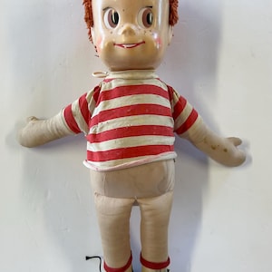 May include: Vintage doll with a painted face, red hair, and a red and white striped shirt. The doll has fabric arms and legs, and is wearing beige shoes with black soles. A classic toy.