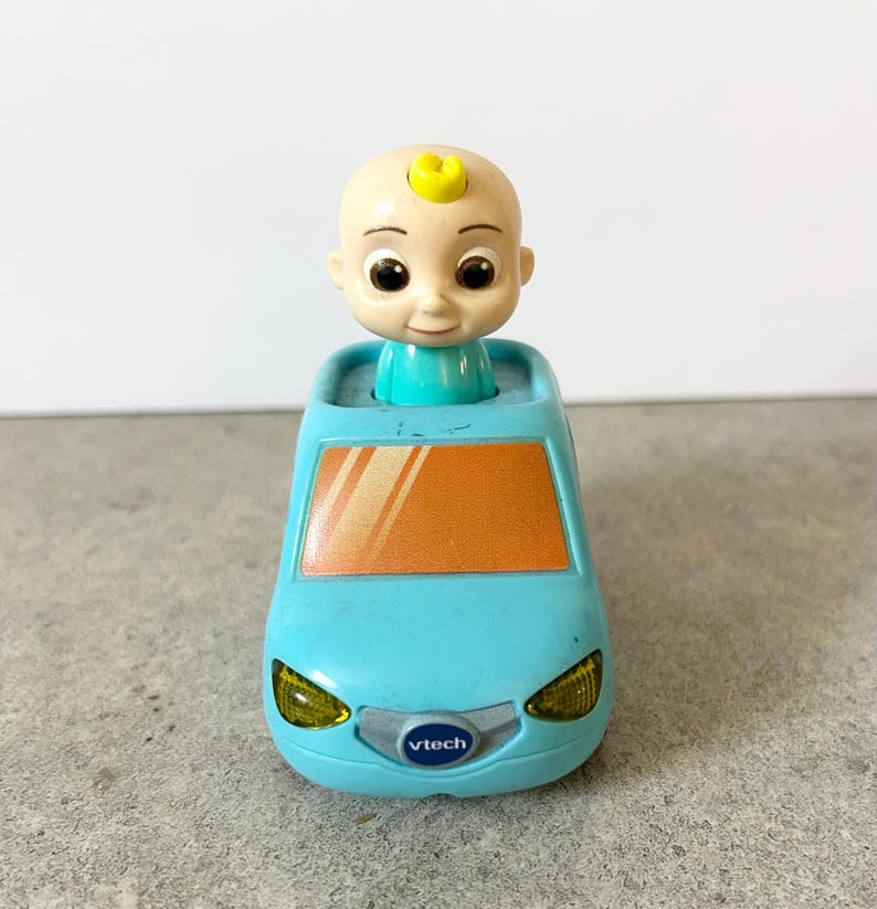 May include: A light blue toy car featuring a cartoon baby figure with a small yellow curl on its head. The car has an orange windshield, yellow headlights, and a silver front grille with the VTech brand logo printed in blue and white text.