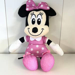 May include: A plush Minnie Mouse doll with a pink polka dot dress, bow, and shoes. The doll has black ears, a white face, and white gloves. The doll is sitting with its legs crossed.