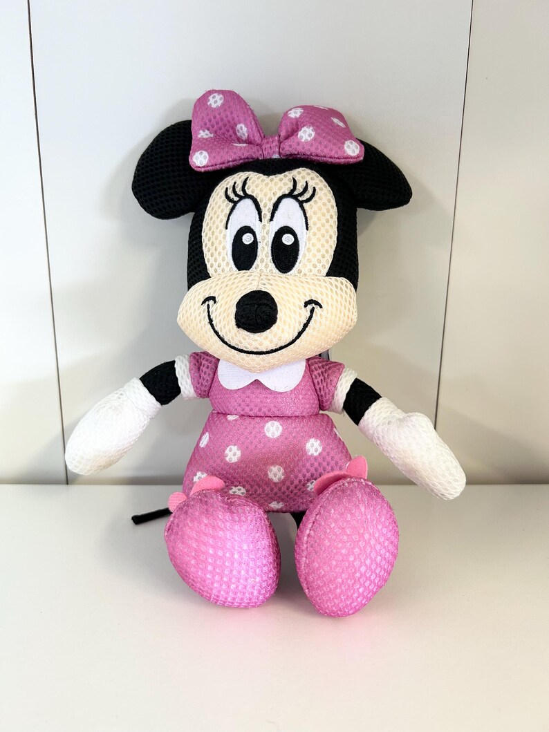 May include: A plush Minnie Mouse doll with a pink polka dot dress, bow, and shoes. The doll has black ears, a white face, and white gloves. The doll is sitting with its legs crossed.