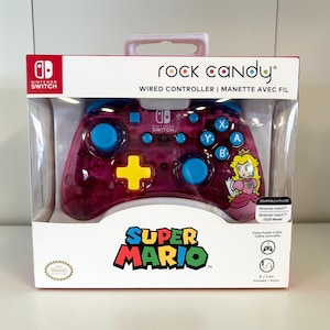 May include: A clear, pink Rock Candy wired controller for Nintendo Switch. The controller has blue buttons and a yellow directional pad. The packaging features the Super Mario logo and Princess Peach.