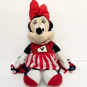 May include: A plush Minnie Mouse doll wearing a red and white cheerleader outfit with a matching bow. The doll has black ears, a black nose, and white gloves. The outfit includes a red top with a white and black graphic and a red and white striped skirt.