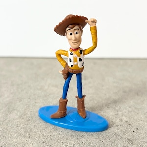 May include: A plastic figurine of a cowboy character wearing a brown hat, a yellow long-sleeve shirt, a white vest with black cow-print spots, blue pants, and brown boots. The figure stands on a blue oval base with one arm raised upward.