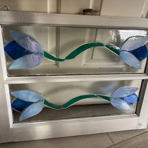 Puede incluir: Un marco de ventana pintado de blanco con dos paneles de vidriera con flores azules y verdes. Las flores son estilizadas y tienen un aspecto caprichoso.