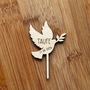 May include: A wooden dove cake topper, featuring the word "TAUFE" and the date "17.04.2024". The dove holds an olive branch. The topper is set against a wooden background.
