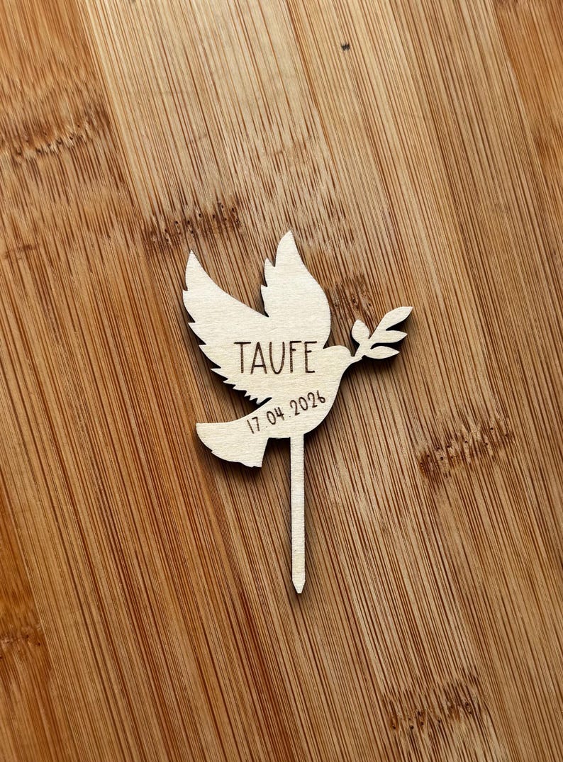 May include: A wooden dove cake topper, featuring the word "TAUFE" and the date "17.04.2024". The dove holds an olive branch. The topper is set against a wooden background.