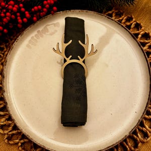 Puede incluir: Un plato de cerámica beige con una servilleta verde oscuro sujeta por un servilletero de madera con forma de cuernos de reno. El plato está sobre un salvamanteles tejido. Bayas rojas y ramas de hoja perenne en el fondo.