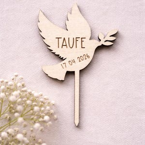 May include: A light brown wooden cake topper in the shape of a dove, with the word "TAUFE" and the date "17.04.2026" engraved. The dove holds an olive branch. The topper has a pointed stick. White flowers are in the lower left corner.