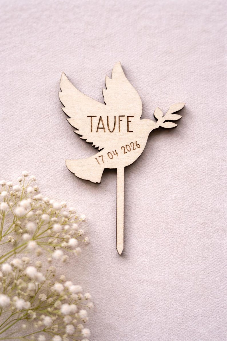 May include: A light brown wooden cake topper in the shape of a dove, with the word "TAUFE" and the date "17.04.2026" engraved. The dove holds an olive branch. The topper has a pointed stick. White flowers are in the lower left corner.