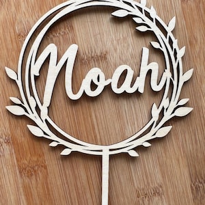 May include: A wooden cake topper featuring the name "Noah" in cursive, surrounded by a wreath of leaves. The light-coloured wood topper has a stem for insertion into a cake. The design is simple and elegant.