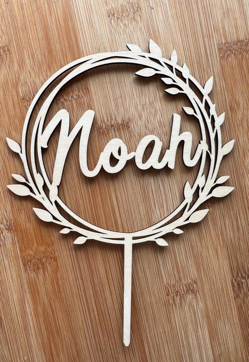 May include: A wooden cake topper featuring the name "Noah" in cursive, surrounded by a wreath of leaves. The light-coloured wood topper has a stem for insertion into a cake. The design is simple and elegant.