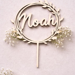 May include: Wooden cake topper with the name "Noah" in a cursive font, encircled by a wreath of leaves. The topper is adorned with small white flowers and has a stake for insertion into a cake.