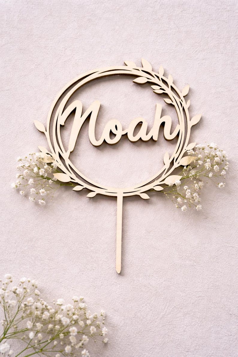 May include: Wooden cake topper with the name "Noah" in a cursive font, encircled by a wreath of leaves. The topper is adorned with small white flowers and has a stake for insertion into a cake.