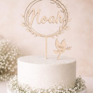 May include: A white cake with a wooden topper displaying the name "Noah" within a wreath. A dove-shaped wooden decoration with the word "TAUFE" and a date is also present. The cake sits on a white stand and is adorned with white flowers.
