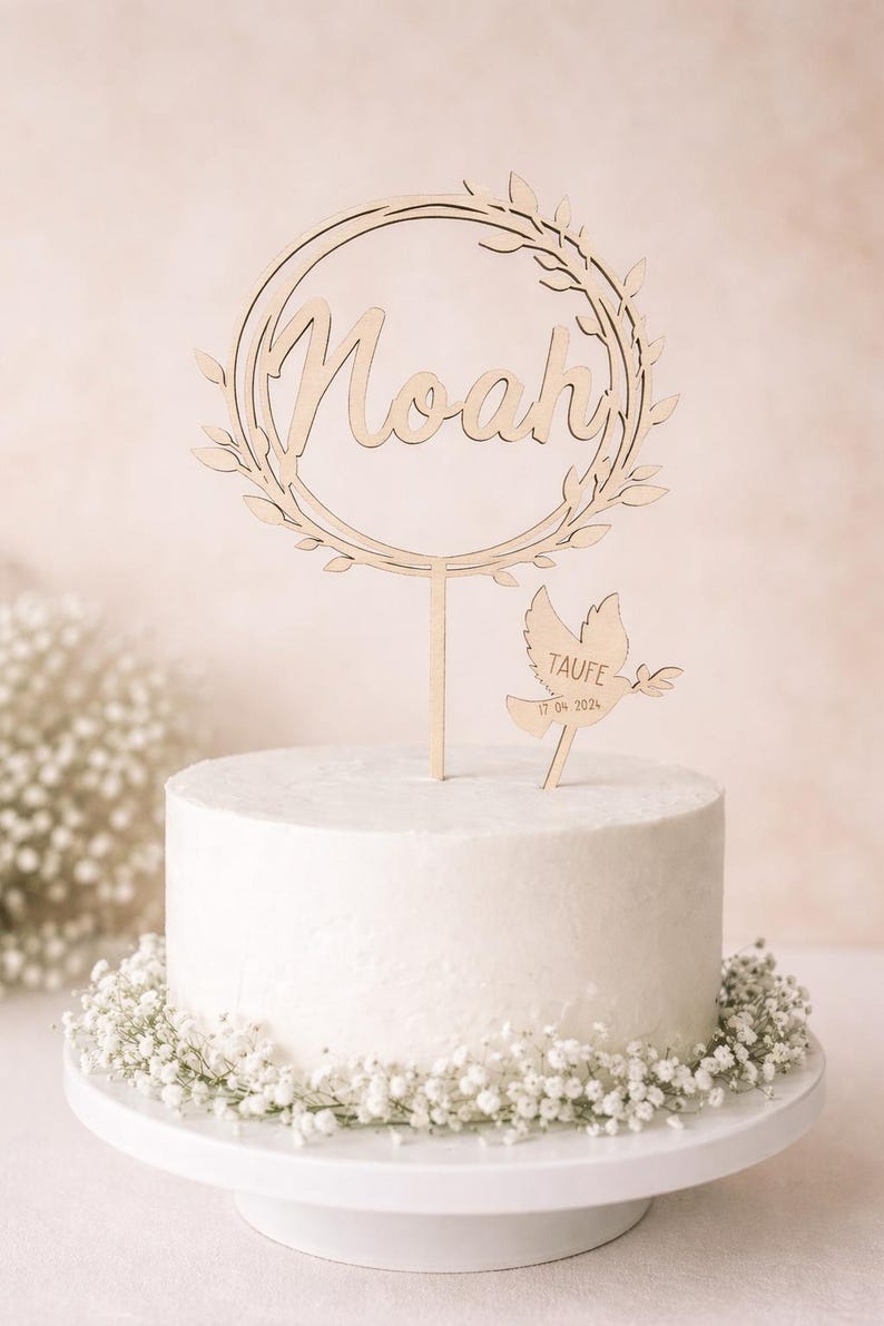 May include: A white cake with a wooden topper displaying the name "Noah" within a wreath. A dove-shaped wooden decoration with the word "TAUFE" and a date is also present. The cake sits on a white stand and is adorned with white flowers.