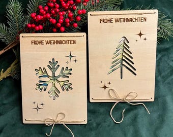 May include: Two wooden Christmas cards with the text "FROHE WEIHNACHTEN". One card features a snowflake cutout, the other a Christmas tree. Both cards have star accents and twine bows.
