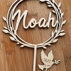 May include: A wooden cake topper featuring the name "Noah" in cursive within a circular wreath. A separate dove-shaped piece reads "Taufe" and "17 04 2024". The topper is made of light-coloured wood.
