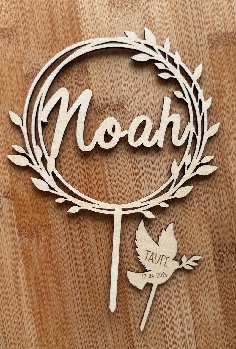 May include: A wooden cake topper featuring the name "Noah" in cursive within a circular wreath. A separate dove-shaped piece reads "Taufe" and "17 04 2024". The topper is made of light-coloured wood.