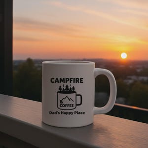 Könnte beinhalten: Eine weiße Keramiktasse mit den Wörtern "CAMPFIRE & COFFEE" und "Dad's Happy Place" in Schwarz bedruckt. Die Tasse zeigt eine Grafik von Bäumen, Bergen und einer Kaffeetasse. Der Hintergrund zeigt einen Sonnenuntergang.