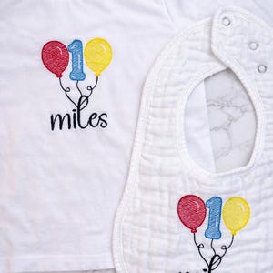 May include: A white short-sleeved t-shirt and a matching white bib, both featuring embroidered designs of a blue number one flanked by a red balloon and a yellow balloon, with the word miles stitched in black cursive script below the balloons.
