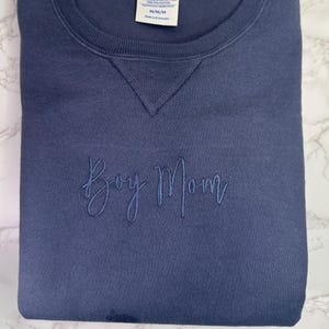May include: A navy blue sweatshirt with the embroidered text "Boy Mom" in a matching shade. The crew neck sweatshirt is folded neatly, showcasing the soft fabric and the delicate script of the embroidered text.
