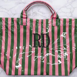 Pode incluir: Uma bolsa tote retangular com acabamento brilhante e listras verticais rosa e verde-escuras. O centro da bolsa exibe as letras de monograma pretas bordadas RD. Inclui duas al&ccedil;as superiores correspondentes com o mesmo padr&atilde;o listrado.