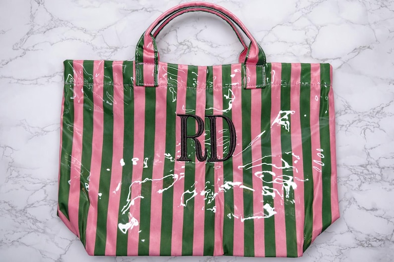 Pode incluir: Uma bolsa tote retangular com acabamento brilhante e listras verticais rosa e verde-escuras. O centro da bolsa exibe as letras de monograma pretas bordadas RD. Inclui duas al&ccedil;as superiores correspondentes com o mesmo padr&atilde;o listrado.