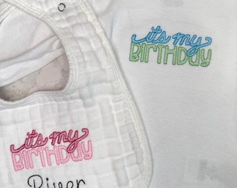 May include: White baby bodysuit and bib set. The bodysuit has the phrase "it's my BIRTHDAY" embroidered in blue and green. The bib has the same phrase embroidered in pink, with the name "River" below it in black.