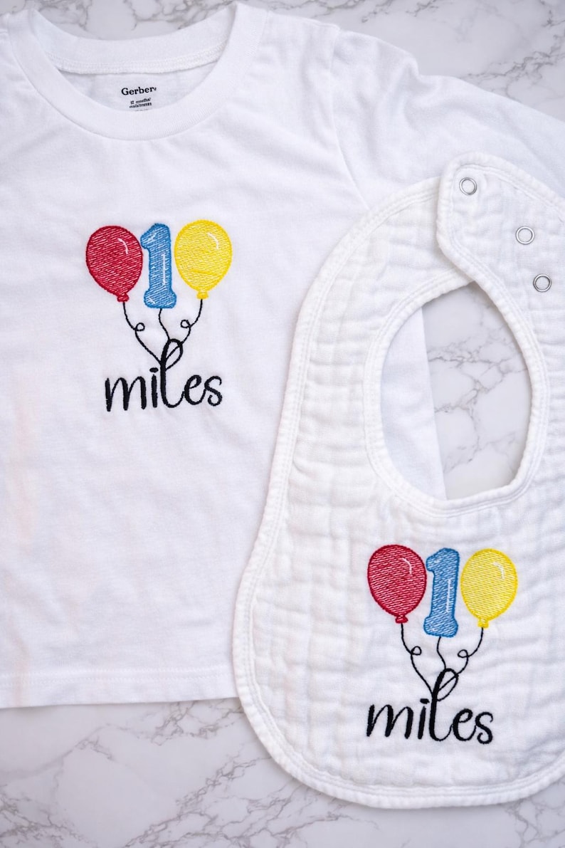 May include: A white short-sleeved t-shirt and a matching white bib, both featuring embroidered designs of a blue number one flanked by a red balloon and a yellow balloon, with the word miles stitched in black cursive script below the balloons.