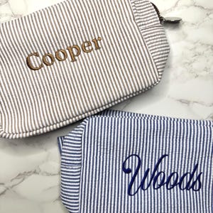 May include: Two striped cosmetic bags. One is brown and white with the name "Cooper" embroidered in a matching brown thread. The other is blue and white with the name "Woods" embroidered in blue thread. Both bags have a zipper closure.