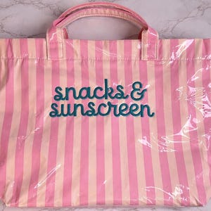 Pode incluir: Uma bolsa tote retangular com listras verticais em tons alternados de rosa claro e p&ecirc;ssego p&aacute;lido. A frente da bolsa exibe o texto snacks & sunscreen bordado em uma fonte cursiva verde-azulado escuro. Possui duas al&ccedil;as superiores.