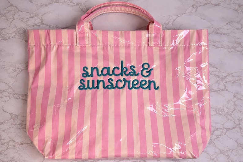 Pode incluir: Uma bolsa tote retangular com listras verticais em tons alternados de rosa claro e p&ecirc;ssego p&aacute;lido. A frente da bolsa exibe o texto snacks & sunscreen bordado em uma fonte cursiva verde-azulado escuro. Possui duas al&ccedil;as superiores.