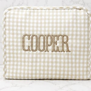May include: A beige and white gingham patterned case with the name "COOPER" embroidered in a brown thread. The case has rounded corners and a soft, quilted appearance. The background is a marble surface.